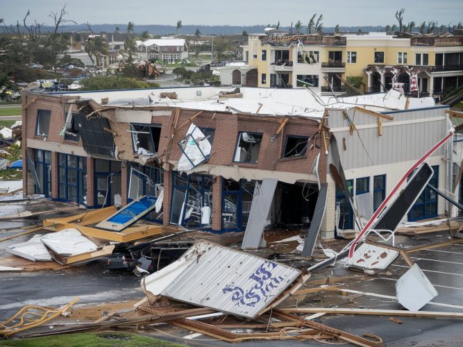 Commercial building severely damaged by hurricane, with debris scattered across parking lot and damaged structures visible in background